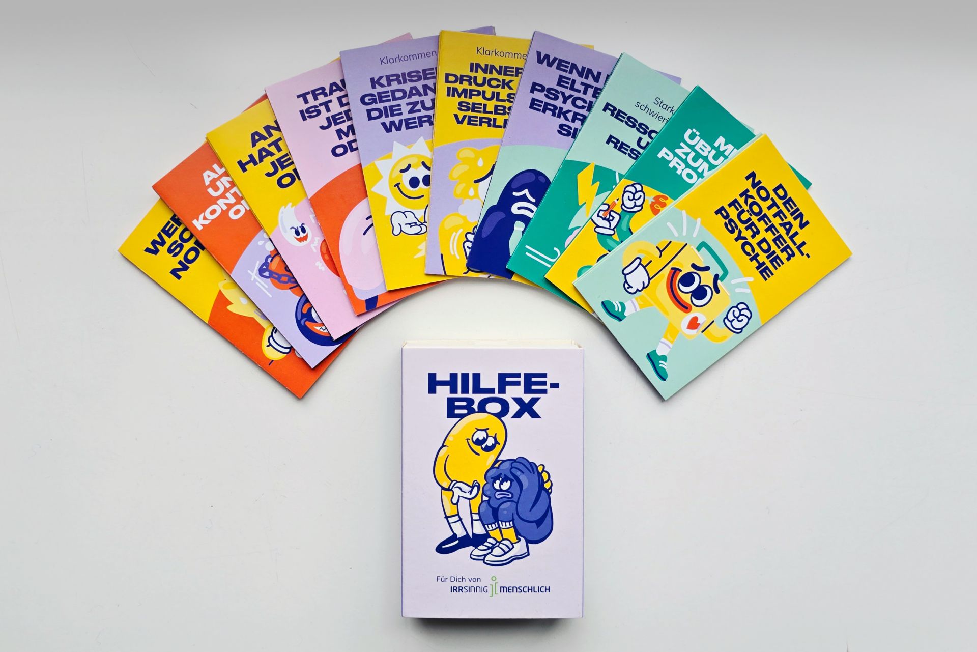 Die neu gestalteten Info-Pocket-Guides von Irrsinnig Menschlich e.V. liegen aufgefächert nebeneinander; die zehn verschiedenen Titel rund um psychische Gesundheit sind sichtbar. Darunter liegt die zugehörige neu gestaltete Hilfebox.