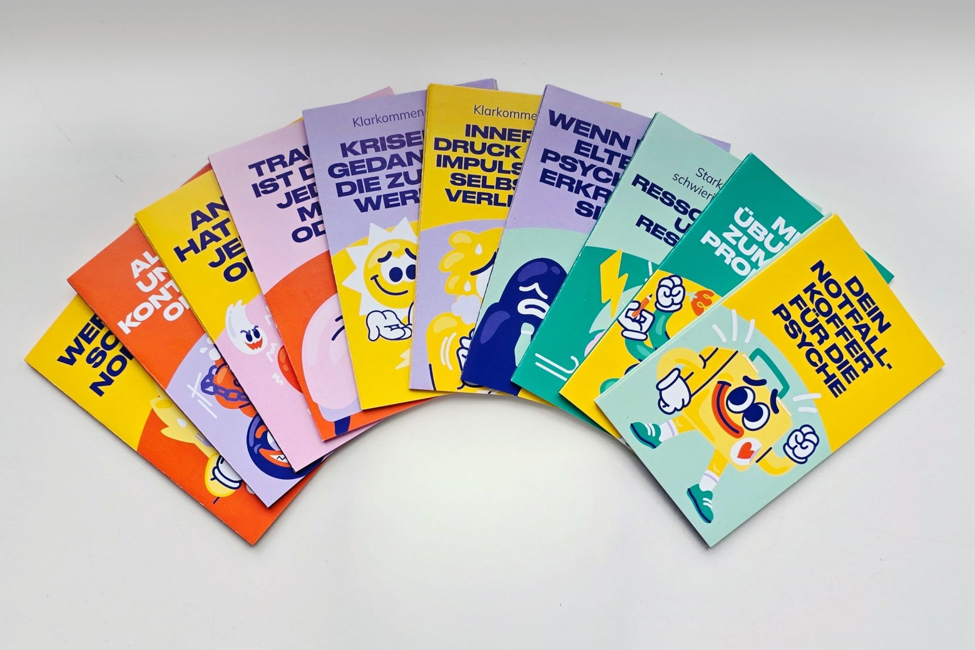 Die neu gestalteten Info-Pocket-Guides von Irrsinnig Menschlich e.V. liegen aufgefächert nebeneinander; die zehn verschiedenen Titel rund um psychische Gesundheit sind sichtbar.